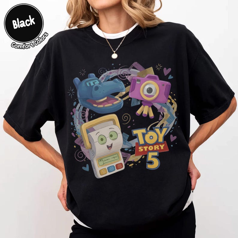 May include: A black t-shirt featuring a graphic print of a blue hippo character, a purple camera, and a white handheld electronic device. The text "TOY STORY 5" is printed in yellow and red. A circular badge in the corner reads "Black Comfort Colors".