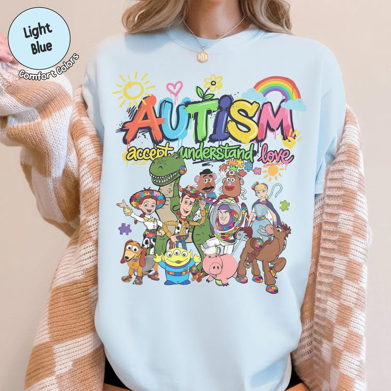K&ouml;nnte beinhalten: Hellblaues T-Shirt mit einem farbenfrohen Grafikdesign. Das Wort "AUTISMUS" ist im Graffiti-Stil dargestellt, begleitet von einem Regenbogen und Zeichentrickfiguren. Der Satz "akzeptieren verstehen lieben" steht unter dem Wort "AUTISMUS".