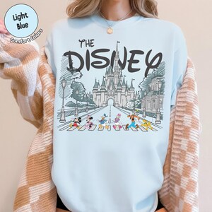 May include: A light blue crewneck sweatshirt featuring a graphic print of a castle and the text THE DISNEY. Below the castle, Mickey Mouse, Minnie Mouse, Donald Duck, Daisy Duck, Goofy, and Pluto are walking across a street in a line.