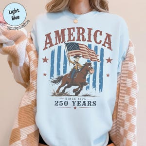 May include: A light blue crewneck sweatshirt featuring a graphic print of a cowboy riding a horse while holding an American flag. The text reads AMERICA, SINCE 1776, and 250 YEARS. The design includes red stars and a distressed blue striped background.