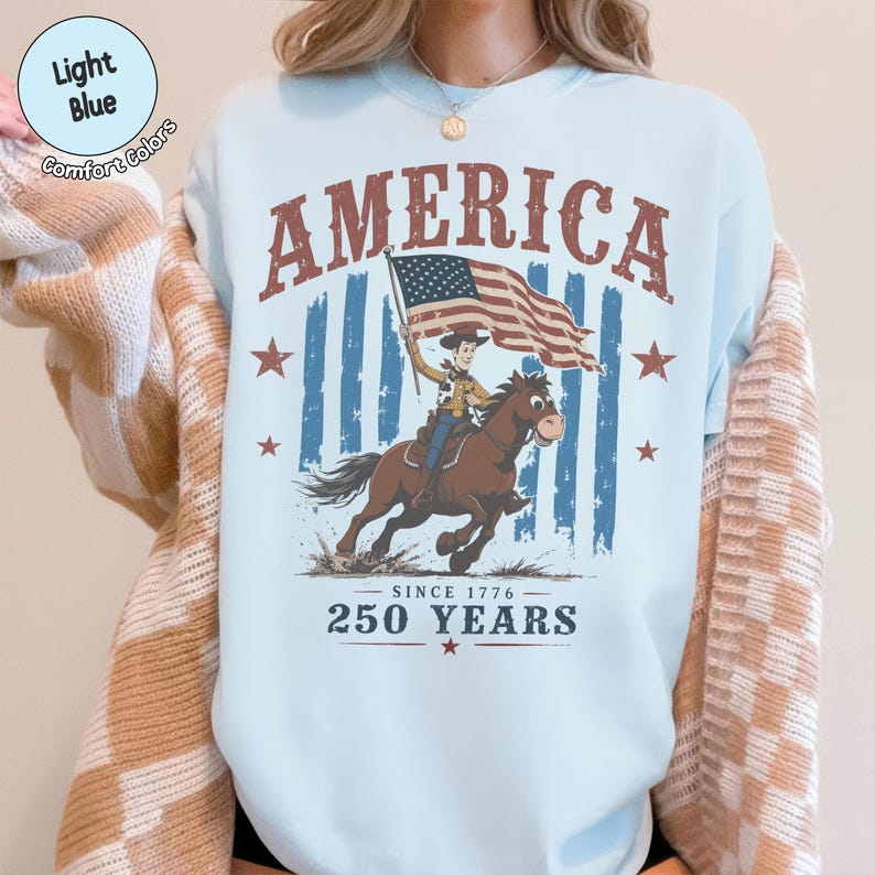 May include: A light blue crewneck sweatshirt featuring a graphic print of a cowboy riding a horse while holding an American flag. The text reads AMERICA, SINCE 1776, and 250 YEARS. The design includes red stars and a distressed blue striped background.