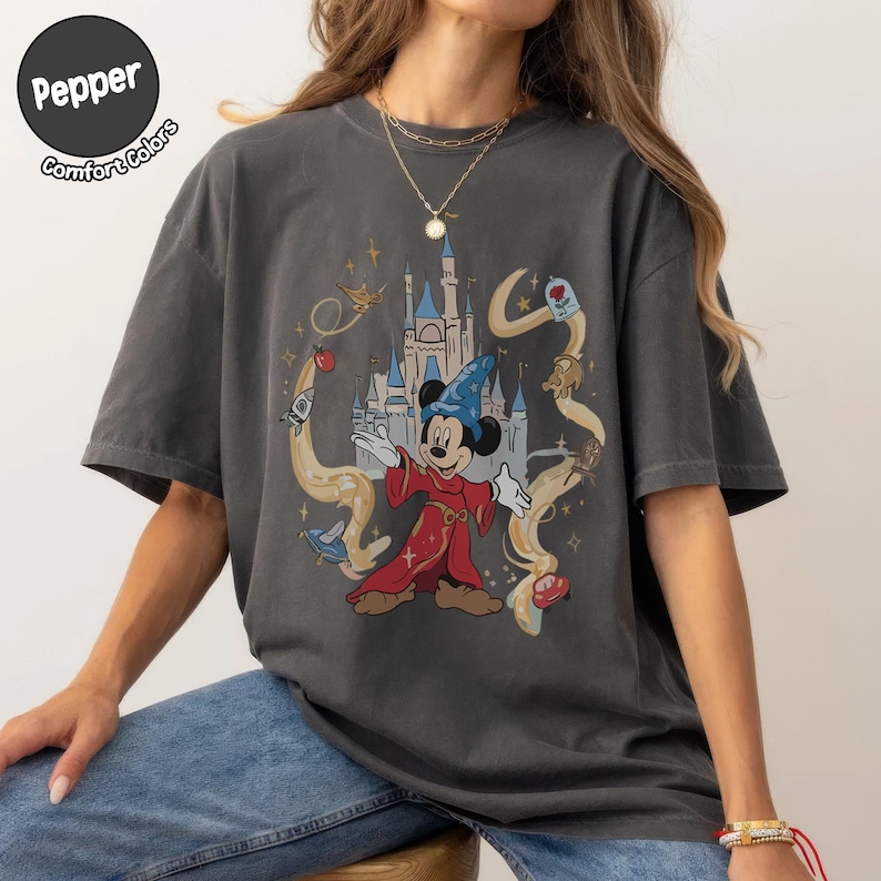 May include: Dark grey t-shirt with a graphic of Mickey Mouse in a red robe and blue wizard hat, with a castle backdrop. The design includes swirling lines and animated elements. The shirt has the text "Pepper Comfort Colors" in a circle.