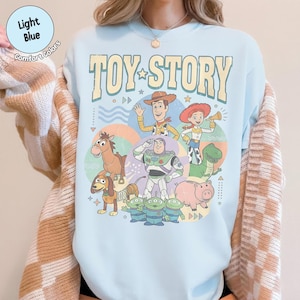 May include: Pale blue "Toy Story" graphic t-shirt featuring characters like Woody and Buzz Lightyear. The shirt displays the words "Toy Story" in a vintage style font. The t-shirt is made of a soft material and is a comfort colors brand.