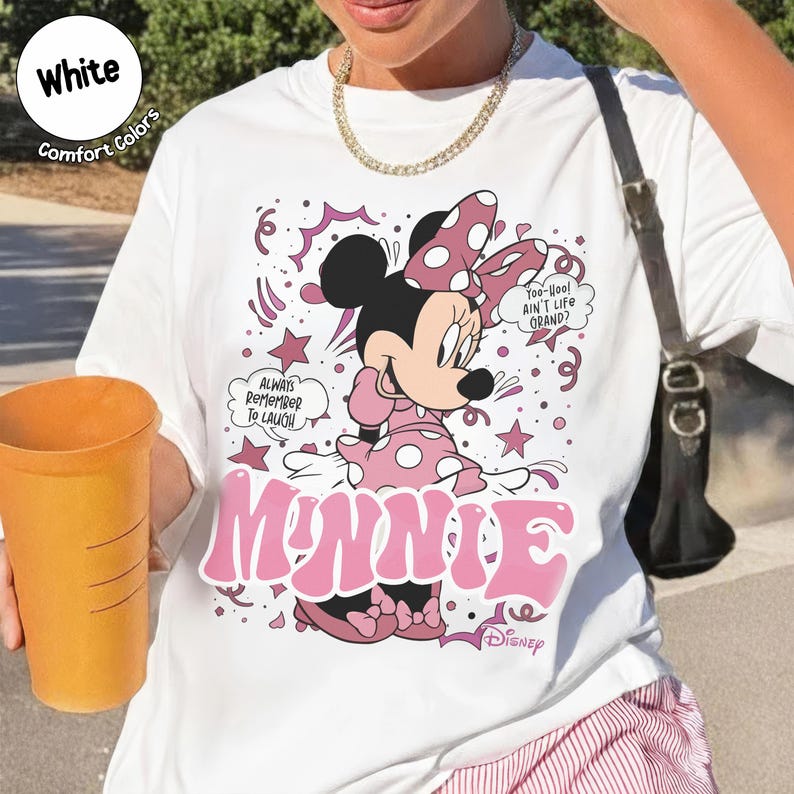May include: White t-shirt featuring Minnie Mouse in a pink polka-dot dress and bow. The design includes the word "MINNIE" in pink, speech bubbles with text, and the Disney logo. The shirt is labeled "Comfort Colors."