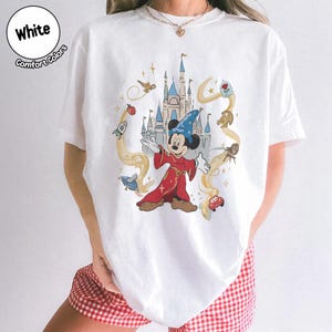 May include: White t-shirt with a vibrant graphic of Mickey Mouse as a sorcerer, surrounded by animated imagery and a castle. The text "White Comfort Colors" is in a circle at the top left.