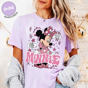 May include: Orchid Comfort Colors t-shirt featuring Minnie Mouse in a pink polka-dot dress and bow. The shirt has the word "MINNIE" in pink, along with the phrases "Always Remember to Laugh" and "Too-Woo! Ain't Life Grand?"