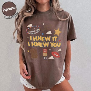 Puede incluir: Una camiseta marr&oacute;n de manga corta con un estampado gr&aacute;fico que incluye el texto I KNEW IT I KNEW YOU, una pulsera de cuentas con el texto YOU'VE GOT A FRIEND IN ME, una bota vaquera marr&oacute;n, un sombrero rojo y bloques apilados con T, S y 5.