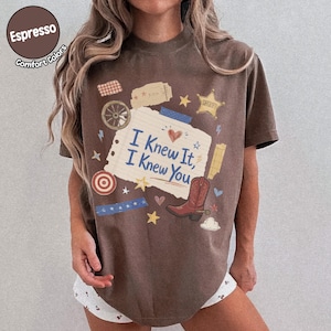 May include: An espresso-colored t-shirt featuring a central graphic of a lined paper scrap with the text I Knew It, I Knew You. The design includes a red cowboy boot, a sheriff star, a wagon wheel, a target, and various small decorative icons.
