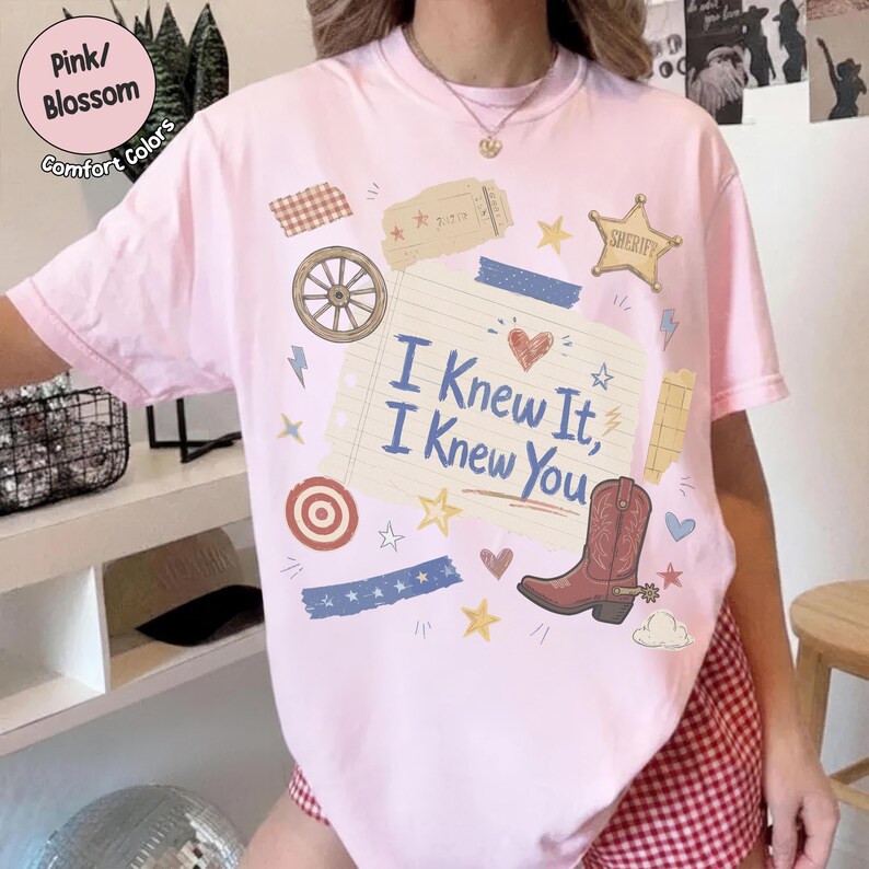 May include: A light pink t-shirt featuring a central graphic of a lined notepad page with the text I Knew It, I Knew You in blue. The design includes a brown cowboy boot, a wagon wheel, a sheriff star, hearts, stars, and decorative tape strips.