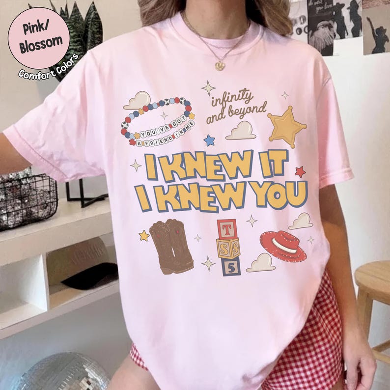 Puede incluir: Una camiseta de manga corta de color rosa claro con un estampado gr&aacute;fico que incluye el texto "I KNEW IT I KNEW YOU", "infinity and beyond" y "YOU'VE GOT A FRIEND IN ME". El dise&ntilde;o incluye ilustraciones de una pulsera de cuentas, una estrella, botas vaqueras, bloques y un sombrero.