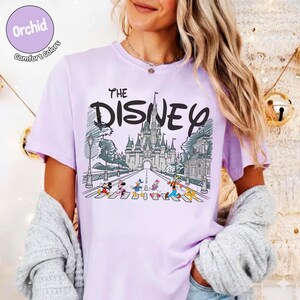 May include: A light purple t-shirt featuring a graphic print of a castle and cartoon characters walking on a crosswalk. The text reads THE DISNEY above the castle illustration. The shirt has a round neckline and short sleeves, with a circular badge in the top left corner that says Orchid Comfort Colors.