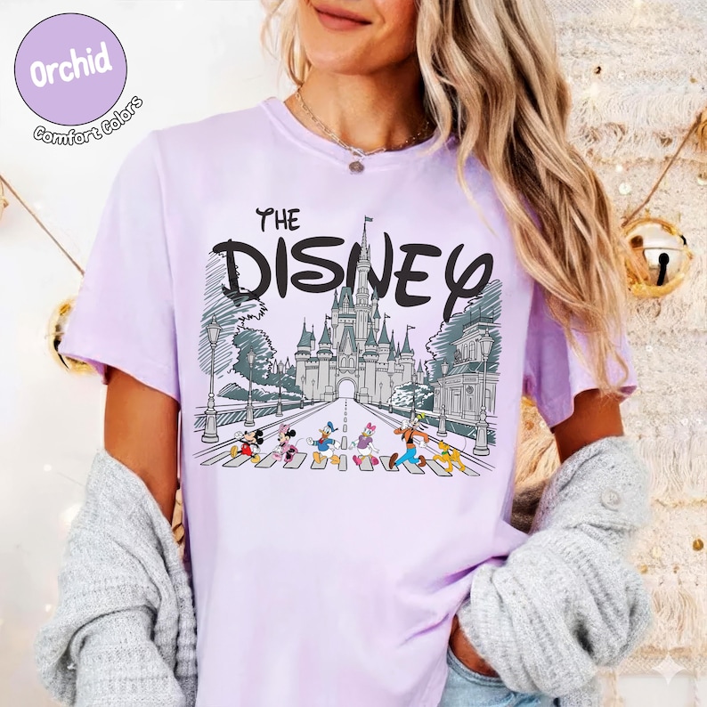 May include: A light purple t-shirt featuring a graphic print of a castle and cartoon characters walking on a crosswalk. The text reads THE DISNEY above the castle illustration. The shirt has a round neckline and short sleeves, with a circular badge in the top left corner that says Orchid Comfort Colors.