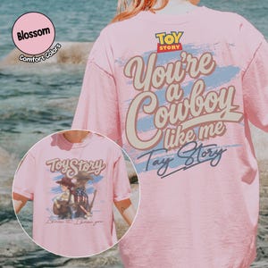 May include: A light pink t-shirt featuring a graphic on the back with the Toy Story logo and the text You're a Cowboy like me Tay Story. The front graphic shows two figures with guitars and the text Toy Story I know it. I know you.