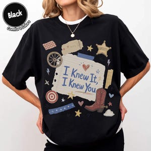 May include: A black t-shirt featuring a central graphic of a lined notebook paper scrap with the text "I Knew It, I Knew You" in blue. The design includes a brown cowboy boot, a sheriff star, a wagon wheel, a heart, and various small stars.