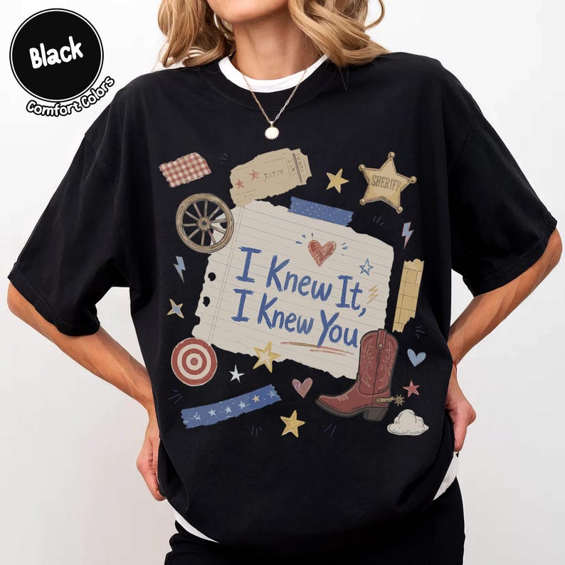 May include: A black t-shirt featuring a central graphic of a lined notebook paper scrap with the text "I Knew It, I Knew You" in blue. The design includes a brown cowboy boot, a sheriff star, a wagon wheel, a heart, and various small stars.