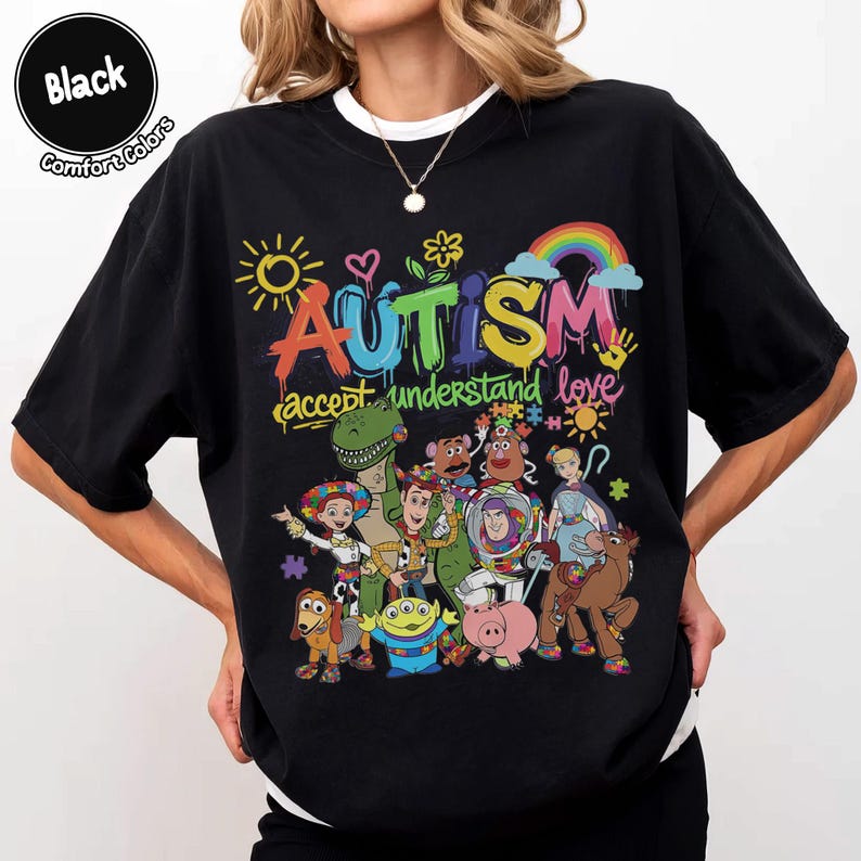 K&ouml;nnte beinhalten: Schwarzes T-Shirt mit einer farbenfrohen Grafik mit dem Wort "AUTISMUS" &uuml;ber einer Sammlung von Zeichentrickfiguren. Das Shirt enth&auml;lt auch die Worte "akzeptieren verstehen lieben" in einer Graffiti-Schriftart.