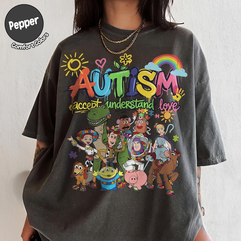 K&ouml;nnte beinhalten: Dunkelgraues T-Shirt mit einem farbenfrohen Aufdruck mit dem Wort "AUTISMUS" und dem Satz "akzeptieren verstehen lieben". Das Design umfasst Zeichentrickfiguren und einen Regenbogen. Das Shirt ist von Pepper Comfort Colors.