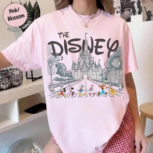 May include: A light pink short-sleeve t-shirt featuring a graphic print of a castle and Disney characters including Mickey, Minnie, Donald, Daisy, Goofy, and Pluto. The text reads THE DISNEY above the castle illustration in a dark font.