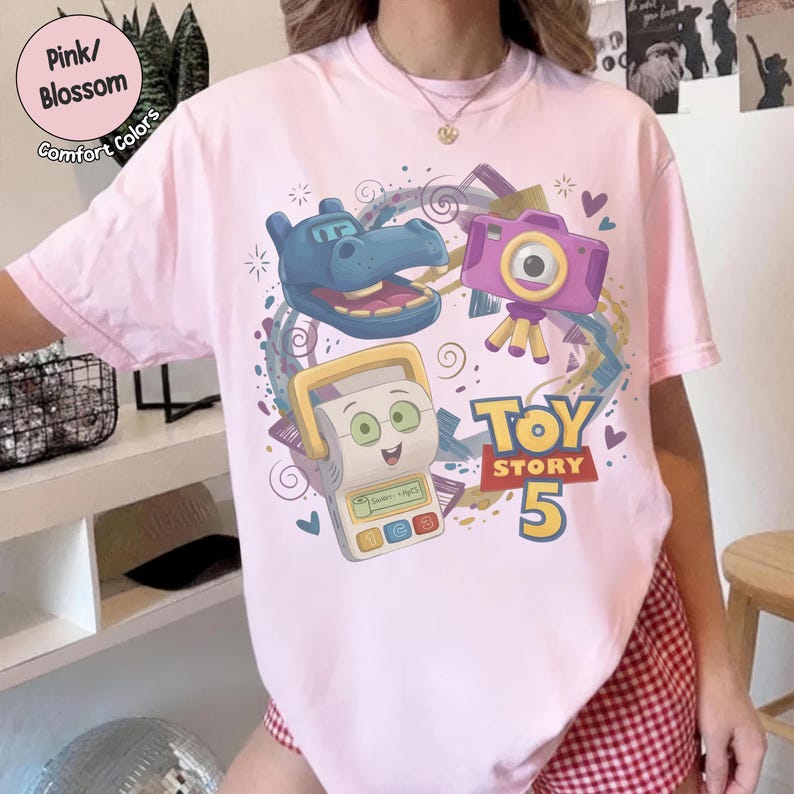 May include: A light pink t-shirt featuring a graphic print of a blue hippo character, a purple camera, and a white toy device. The design includes the text "TOY STORY 5" in yellow and red, surrounded by colorful swirls, small hearts, and star shapes.
