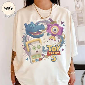 May include: An ivory t-shirt featuring a graphic print with a blue hippo character, a purple camera on a tripod, and a white handheld electronic game device. The text Toy Story 5 is printed in bold yellow and red letters, surrounded by purple swirls.
