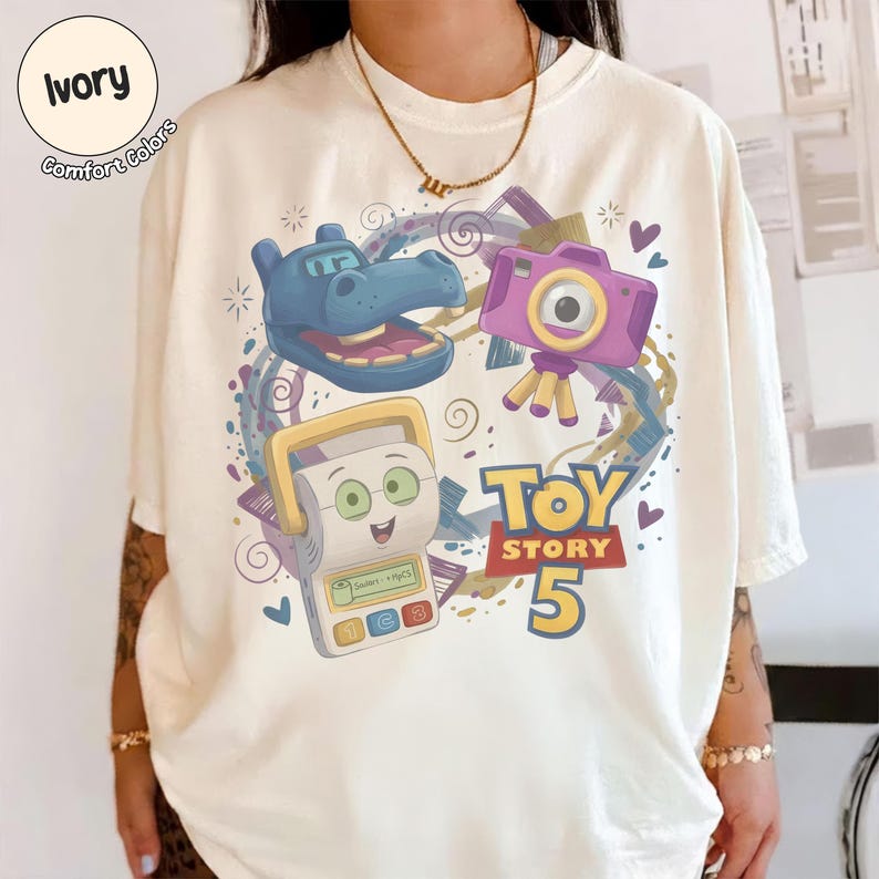 May include: An ivory t-shirt featuring a graphic print with a blue hippo character, a purple camera on a tripod, and a white handheld electronic game device. The text Toy Story 5 is printed in bold yellow and red letters, surrounded by purple swirls.