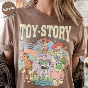 May include: Espresso-coloured t-shirt with the words "TOY STORY" in a vintage font, and cartoon characters including Woody and Buzz Lightyear. The shirt has a crew neck and short sleeves. The words "Comfort Colors" are also printed on the shirt.