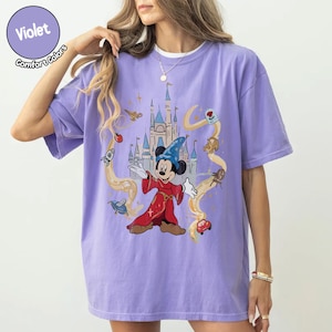 May include: A lilac Comfort Colors t-shirt with a graphic of Mickey Mouse as a sorcerer in front of a castle. The design includes animated elements in yellow and red. The shirt has a crew neck.