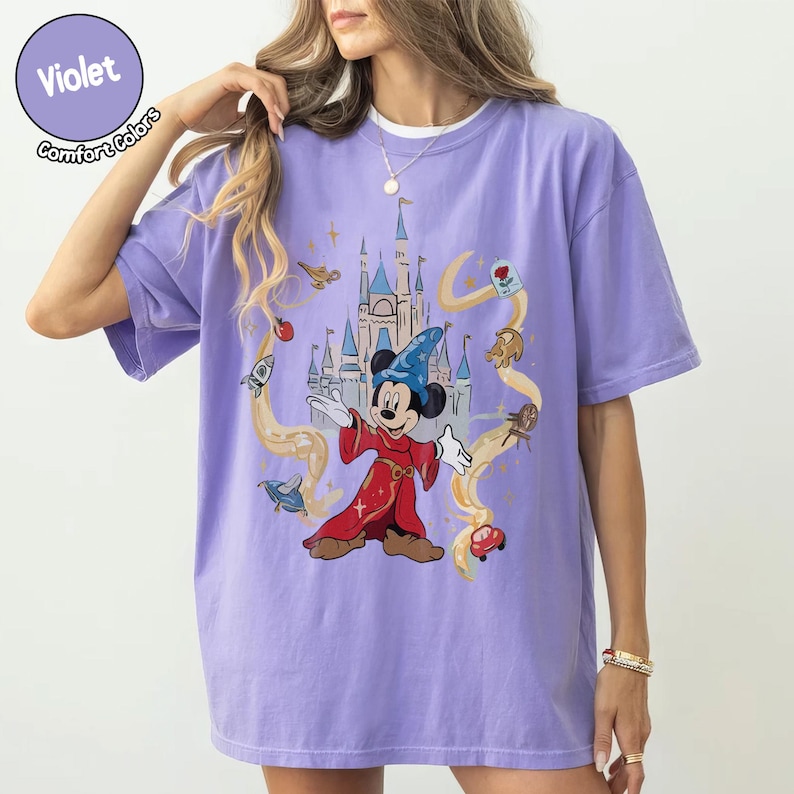 May include: A lilac Comfort Colors t-shirt with a graphic of Mickey Mouse as a sorcerer in front of a castle. The design includes animated elements in yellow and red. The shirt has a crew neck.