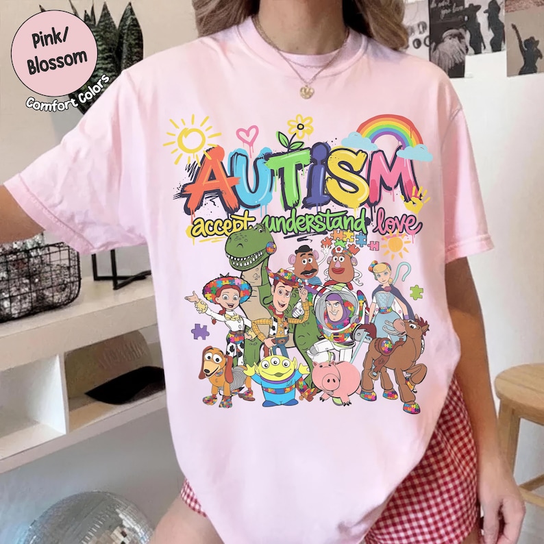 K&ouml;nnte beinhalten: Hellrosa T-Shirt mit dem Wort "AUTISMUS" in bunten Buchstaben, begleitet von den S&auml;tzen "akzeptieren, verstehen, lieben" und Zeichentrickfiguren. Das Shirt zeigt auch einen Regenbogen, eine Sonne und Blumen.