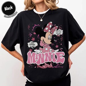 May include: Black t-shirt featuring Minnie Mouse in a pink polka-dot dress and bow. The design includes the word "MINNIE" in pink bubble letters, speech bubbles, and the text "Always Remember To Laugh".