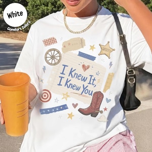 May include: A white t-shirt featuring a graphic print of a lined notebook paper with the text I Knew It, I Knew You, surrounded by western-themed illustrations including a cowboy boot, a sheriff star, a wagon wheel, and various small hearts and stars.