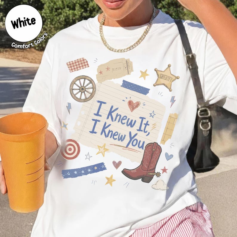 May include: A white t-shirt featuring a graphic print of a lined notebook paper with the text I Knew It, I Knew You, surrounded by western-themed illustrations including a cowboy boot, a sheriff star, a wagon wheel, and various small hearts and stars.
