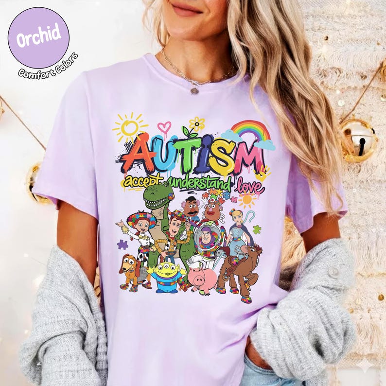 K&ouml;nnte beinhalten: Ein helllila T-Shirt mit einem farbenfrohen Aufdruck mit dem Wort "AUTISMUS" und dem Satz "akzeptieren verstehen lieben". Die Grafik enth&auml;lt einen Regenbogen, eine Sonne und verschiedene Zeichentrickfiguren. Das Shirt hat kurze &Auml;rmel.