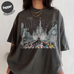 May include: A charcoal gray short-sleeve t-shirt featuring a graphic print of a castle and Disney characters walking on a crosswalk. The text reads THE DISNEY. A circular logo in the top left corner contains the text Pepper Comfort Colors.