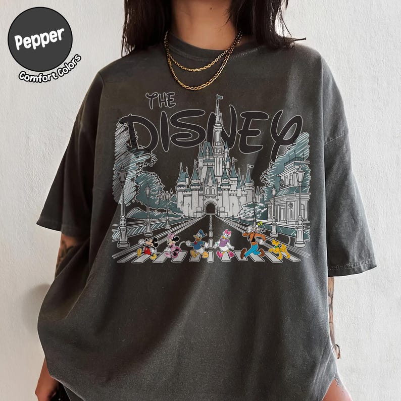 May include: A charcoal gray short-sleeve t-shirt featuring a graphic print of a castle and Disney characters walking on a crosswalk. The text reads THE DISNEY. A circular logo in the top left corner contains the text Pepper Comfort Colors.