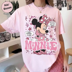 May include: A light pink t-shirt featuring a graphic of Minnie Mouse in a pink polka-dot dress and bow. The word "MINNIE" is printed in large pink letters. Text includes "Always Remember To Laugh" and "Too-Hoo! Ain't Life Grand?"