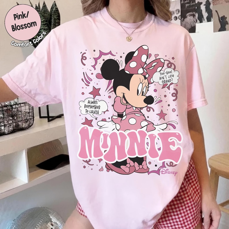 May include: A light pink t-shirt featuring a graphic of Minnie Mouse in a pink polka-dot dress and bow. The word "MINNIE" is printed in large pink letters. Text includes "Always Remember To Laugh" and "Too-Hoo! Ain't Life Grand?"