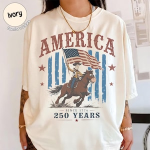 May include: An ivory t-shirt featuring a graphic print with the word AMERICA in red letters at the top. The design shows a cartoon cowboy riding a brown horse while holding an American flag. Below is the text SINCE 1776 250 YEARS with red stars.
