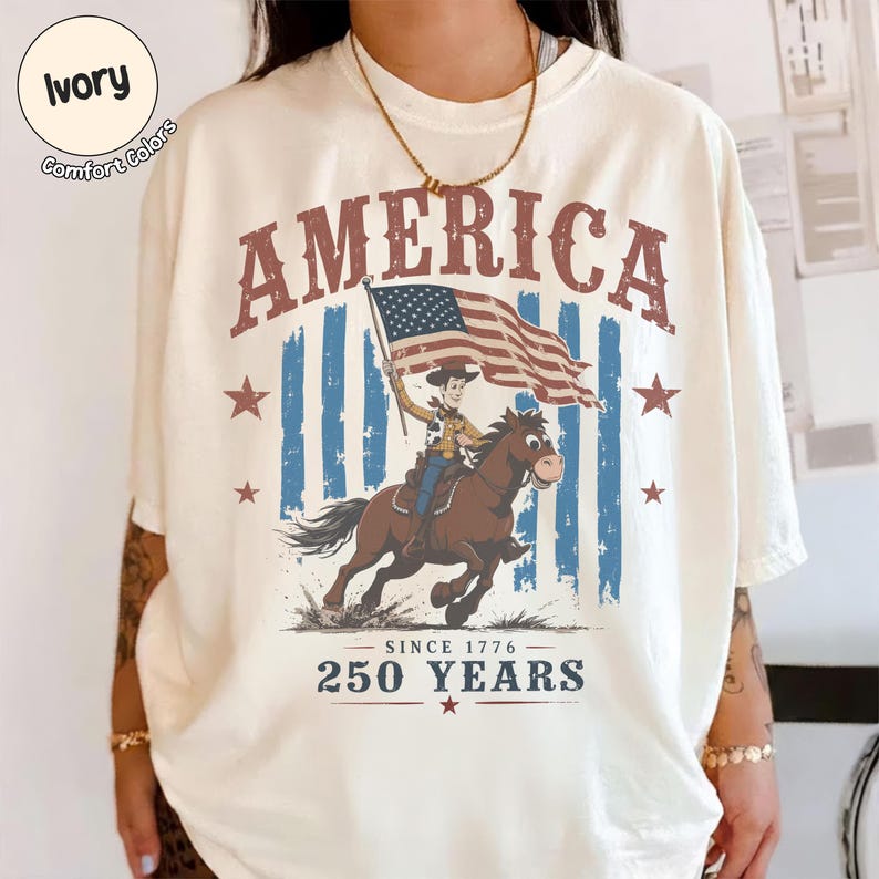 May include: An ivory t-shirt featuring a graphic print with the word AMERICA in red letters at the top. The design shows a cartoon cowboy riding a brown horse while holding an American flag. Below is the text SINCE 1776 250 YEARS with red stars.