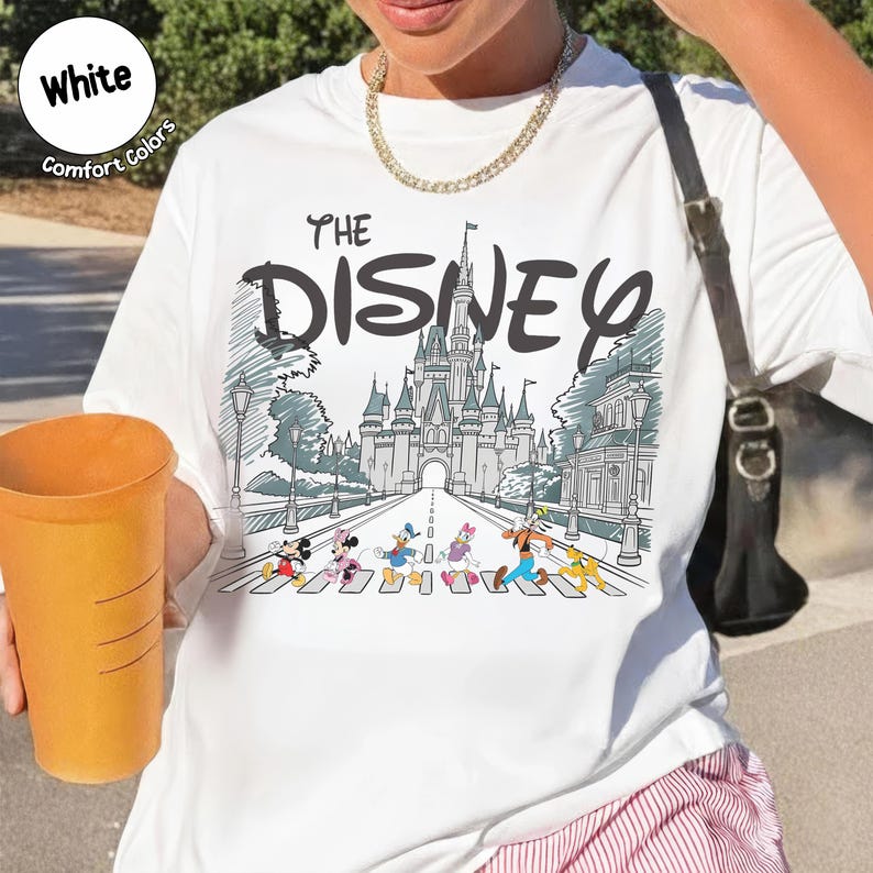 May include: A white t-shirt featuring a graphic print of a castle and cartoon characters walking on a crosswalk. The text reads THE DISNEY. The design includes Mickey Mouse, Minnie Mouse, Donald Duck, Daisy Duck, Goofy, and Pluto in color.