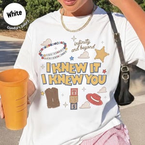 Puede incluir: Una camiseta blanca con un estampado gr&aacute;fico con el texto I KNEW IT I KNEW YOU, infinity and beyond, y YOU'VE GOT A FRIEND IN ME. El dise&ntilde;o incluye ilustraciones de una pulsera de cuentas, una estrella de sheriff, botas vaqueras y un sombrero.