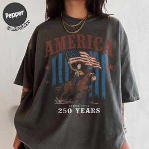 May include: A charcoal gray t-shirt featuring a graphic print with the text AMERICA at the top and SINCE 1776 250 YEARS at the bottom. The design depicts a cartoon cowboy riding a brown horse while holding an American flag, set against blue stripes.