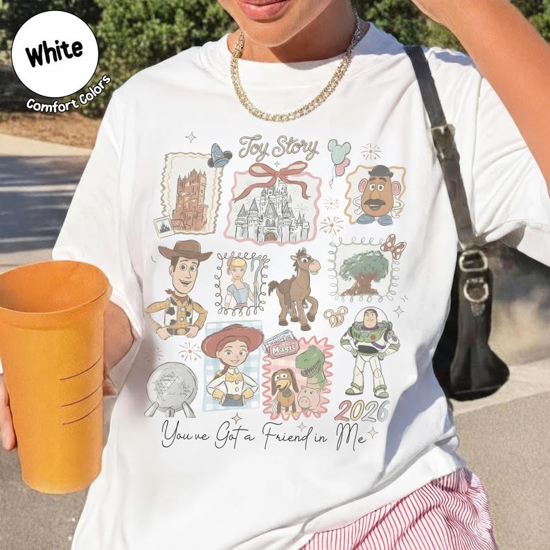 May include: White t-shirt featuring a colorful "Toy Story" design with various characters and the text "You've Got a Friend in Me". The shirt includes the year "2026" and is made of Comfort Colors material.
