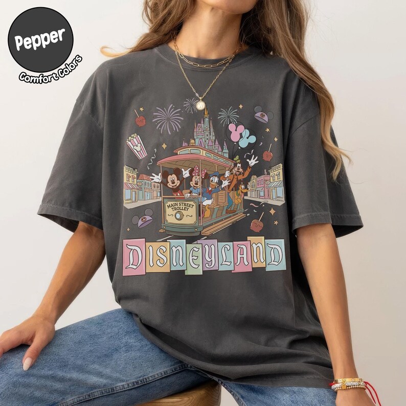 K&ouml;nnte beinhalten: Dunkelgraues T-Shirt mit Disneyland-Design. Die Grafik zeigt eine Stra&szlig;enbahn mit Mickey Mouse, Minnie Mouse, Donald Duck und Goofy, ein Schloss, Feuerwerk und das Wort "Disneyland" in Pastellfarben. Das Shirt hat kurze &Auml;rmel.