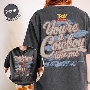 May include: A charcoal gray t-shirt featuring a large graphic on the back with the text "Toy Story" and "You're a Cowboy like me" in a stylized font. The front graphic shows two figures with guitars and the text "Toy Story" and "I knew it, I knew you."