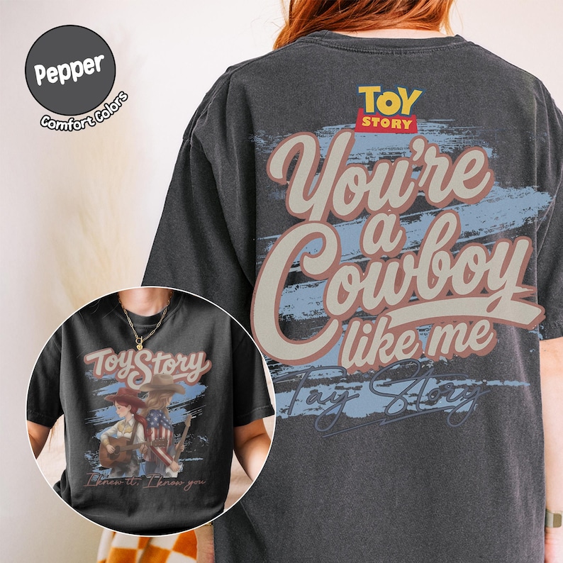 May include: A charcoal gray t-shirt featuring a large graphic on the back with the text "Toy Story" and "You're a Cowboy like me" in a stylized font. The front graphic shows two figures with guitars and the text "Toy Story" and "I knew it, I knew you."