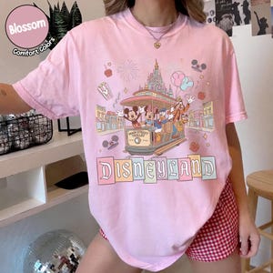 K&ouml;nnte beinhalten: Hellrosa T-Shirt mit Disneyland-Grafik. Das Design zeigt Micky Maus, Minnie Maus, Donald Duck und Goofy in einer Main Street Trolley, mit dem Disneyland-Logo darunter. Auf dem Shirt steht "Blossom Comfort Colors".
