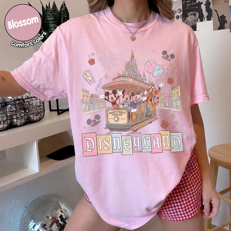 K&ouml;nnte beinhalten: Hellrosa T-Shirt mit Disneyland-Grafik. Das Design zeigt Micky Maus, Minnie Maus, Donald Duck und Goofy in einer Main Street Trolley, mit dem Disneyland-Logo darunter. Auf dem Shirt steht "Blossom Comfort Colors".