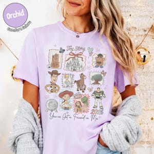 May include: A light purple t-shirt featuring a collage of Toy Story characters and imagery. The design includes illustrations of Woody, Buzz Lightyear, and other characters, along with the text "Toy Story" and "You've Got a Friend in Me."