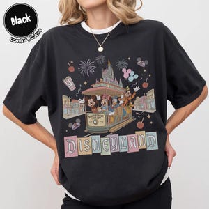 K&ouml;nnte beinhalten: Schwarzes T-Shirt mit einem Disneyland Main Street Trolley-Design. Die Grafik zeigt Micky Maus, Minnie Maus, Donald Duck und Goofy auf der Stra&szlig;enbahn, mit Feuerwerk und dem Schloss im Hintergrund. Das Wort "Disneyland" steht unten.
